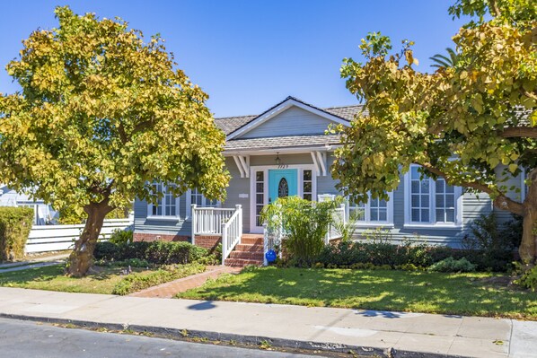 A two-story house with a front porch, a blue door, and a small garden with trees and shrubs.