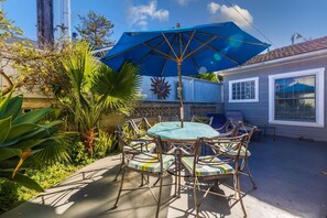 A patio with a round table and chairs, a large blue umbrella, and a house in the background.
