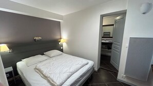Une chambre d’hôtel comprenant un lit, une table de chevet, une lampe, un luminaire fixé au mur, une porte de salle de bain et un système de ventilation.