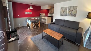 Un salon moderne avec un canapé gris, une table basse en bois et un coin repas agrémenté d’un mur rouge, avec une cuisine en arrière-plan.