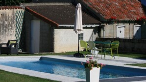 Una piscina con solárium empedrado, un pequeño edificio con tejado de cerámica y una iglesia con campanario al fondo.