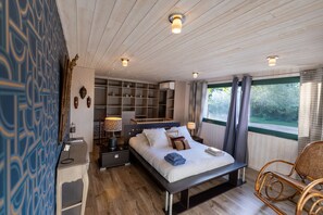 Ein Schlafzimmer mit Bett, Nachttischen, einem Sessel, einem Kleiderschrank und einem großen Fenster mit Vorhängen.