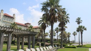 Una fila de sillas blancas bajo una pérgola, palmeras y un edificio con techo rojo.