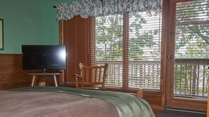 Een slaapkamer met een bed, een televisie, een houten stoel en een balkon met uitzicht op bomen.