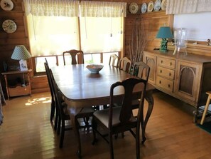 A dining area with a wooden table and chairs, a small side table with a lamp, and a window with blinds.