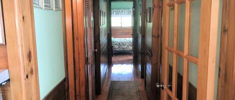 A hallway with wooden doors and a carpeted floor leading to a bedroom with a bed and a window.