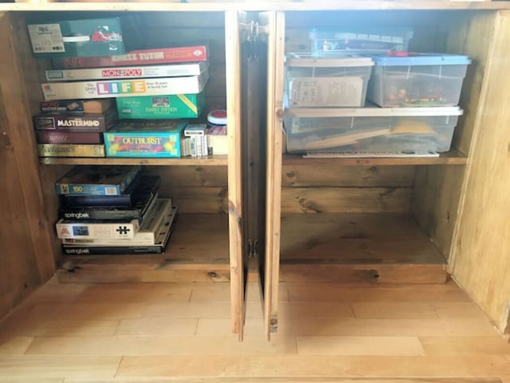 A wooden cabinet with shelves containing board games and storage bins.