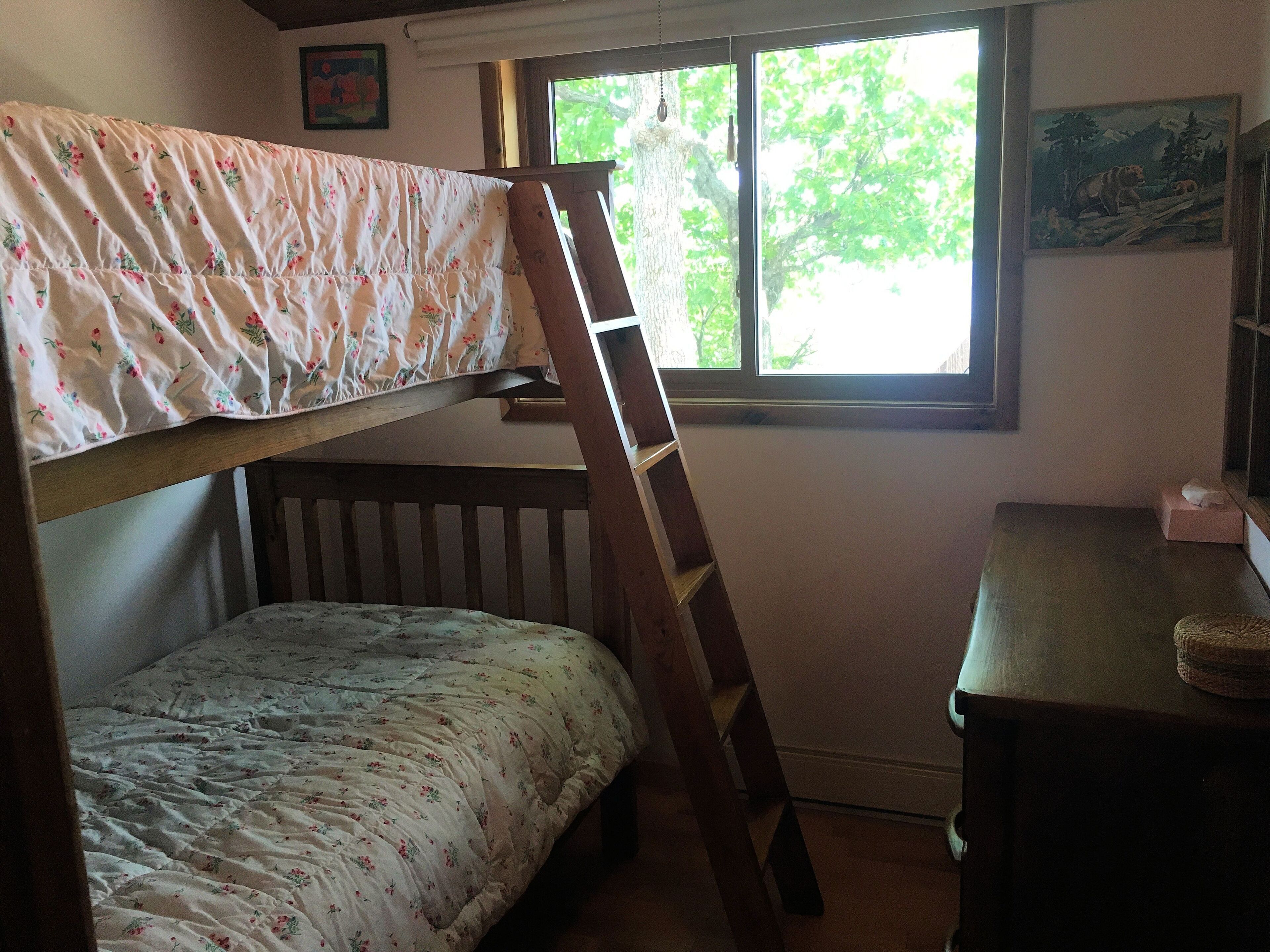 A bunk bed with floral bedding, a wooden ladder, a desk with a chair, and a window with a view of trees.