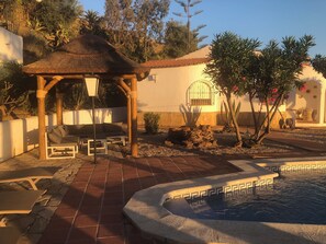 Un espace patio agrémenté d’un gazebo à toit de chaume, de sièges d’extérieur, d’une petite piscine et d’un jardin planté de cactus et d’arbres.