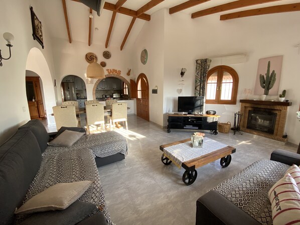 A spacious living room with a sofa, a coffee table, and a television. There is a kitchen area with cabinets and a dining table.
