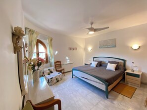 Une chambre à coucher comprenant un lit, des tables de chevet, un bureau, une chaise, un ventilateur de plafond et une fenêtre avec des rideaux.