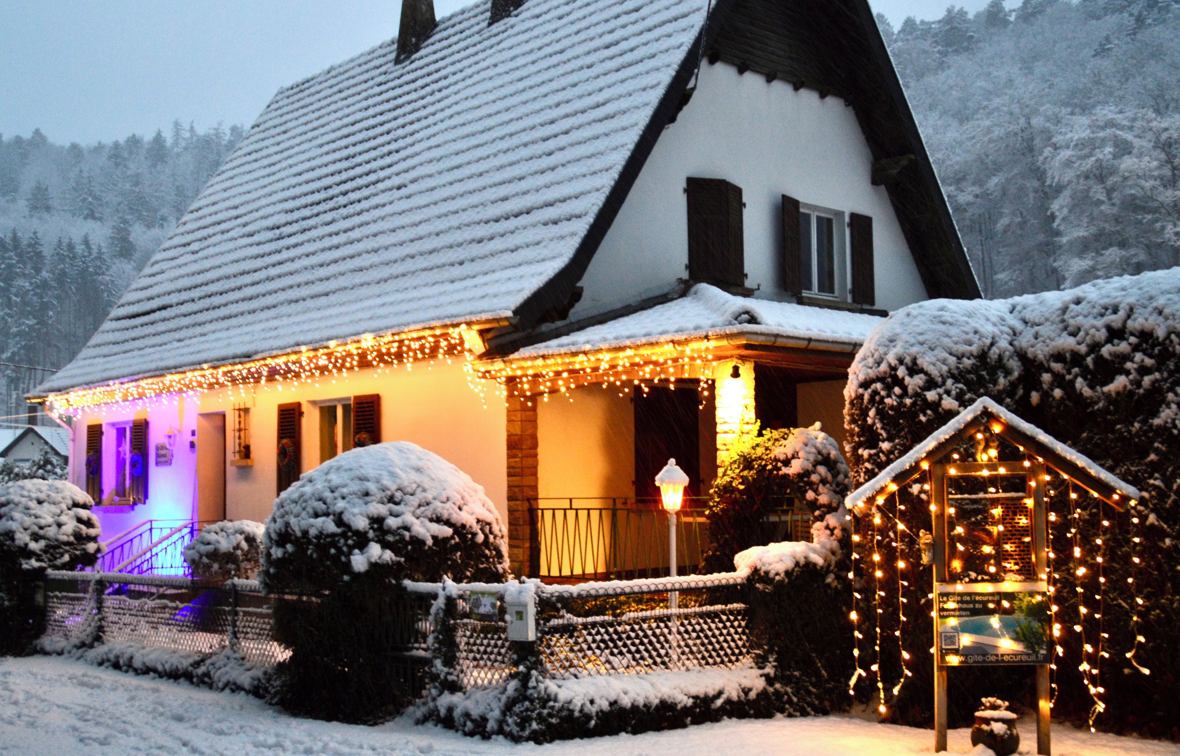 Une maison recouverte de neige, avec un toit en pente, une véranda éclairée et un petit abri de jardin également illuminé.