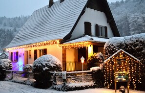 A snow-covered house with a sloped roof, a lit porch, and a small shed with lights. - Comfortable, quiet and independent cottage with swimming pool in the heart (Sturzelbronn)