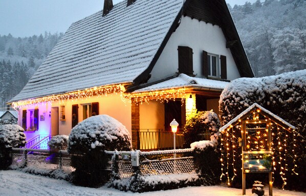 A snow-covered house with a sloped roof, a lit porch, and a small shed with lights.