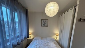 Una cama bien hecha con una colcha blanca y almohadas, dos lámparas de noche, un cuadro en la pared y un ventanal con cortinas traslúcidas.
