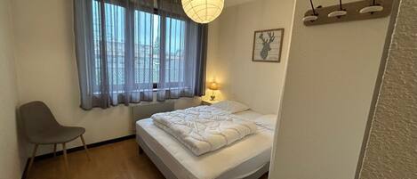 A bedroom with a bed, a chair, a bedside table with a lamp, a window with curtains, and a wall-mounted light fixture.