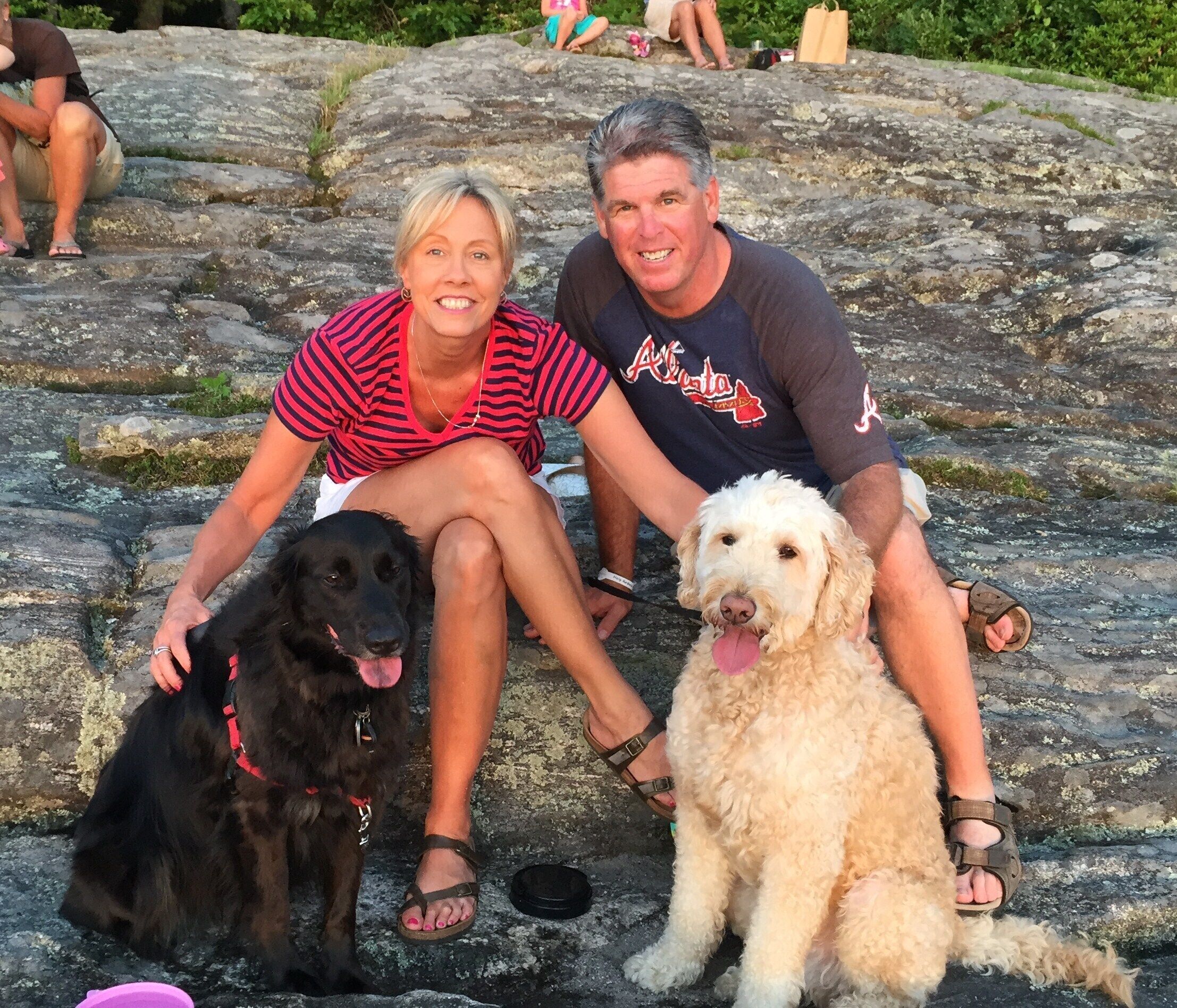 A couple sitting on a rock with two dogs, one black and one white, in a natural setting.