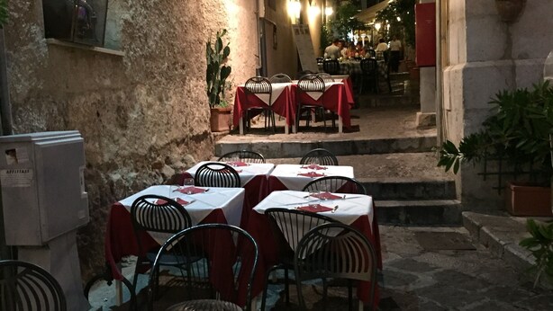 An outdoor dining area with tables and chairs, a staircase leading to an upper level, and a building with a stone facade. - Cefalù Appart 4 (Cefalù)