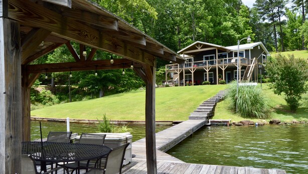 A wooden deck with a table and chairs, a pond, and a house in the background. - Spring special! Enjoy Lake Martin at its best! (Jacksons Gap)