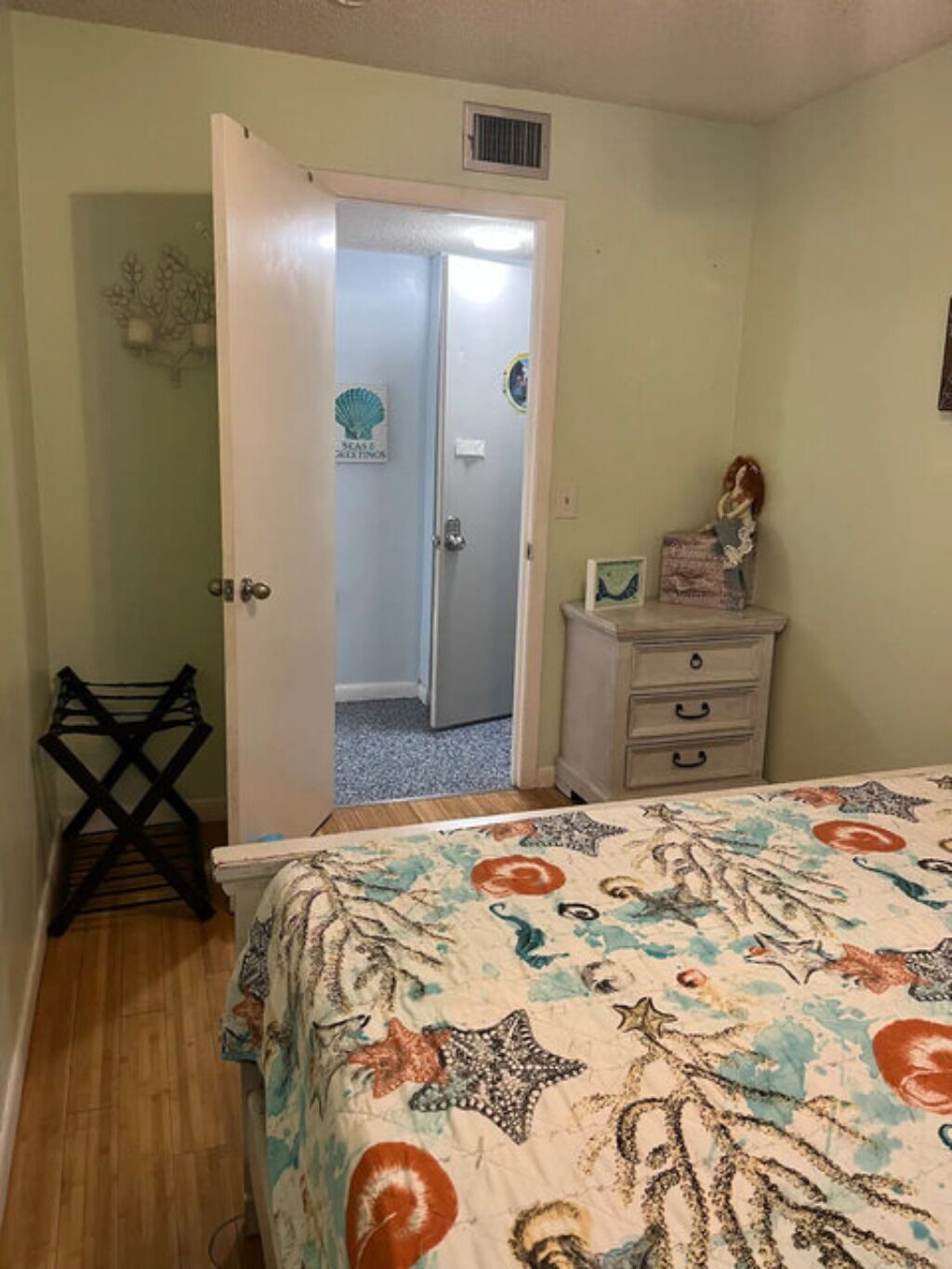 A bedroom with a bed, a dresser, a door leading to another room, and a small shelf with items.