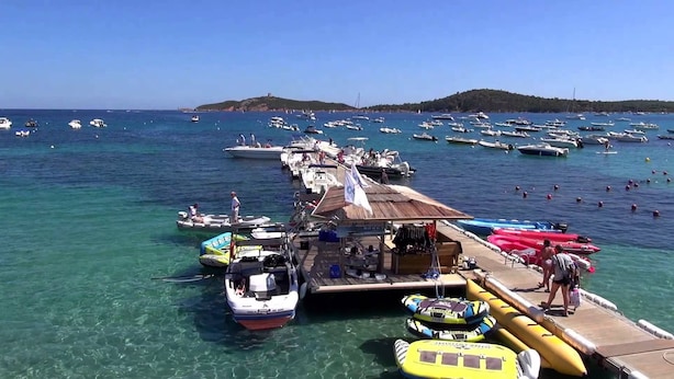 Un port de plaisance où sont amarrés différents types de bateaux, une jetée où se trouvent des personnes et du matériel, et une vue dégagée sur l’eau et les terres environnantes.