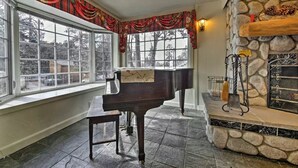 A room with a grand piano, large windows, a stone fireplace, and a stone wall. - Steps to Bear Mountain & Golf Course Spacious 4BR Prime Moonridge Location (Big Bear Lake)