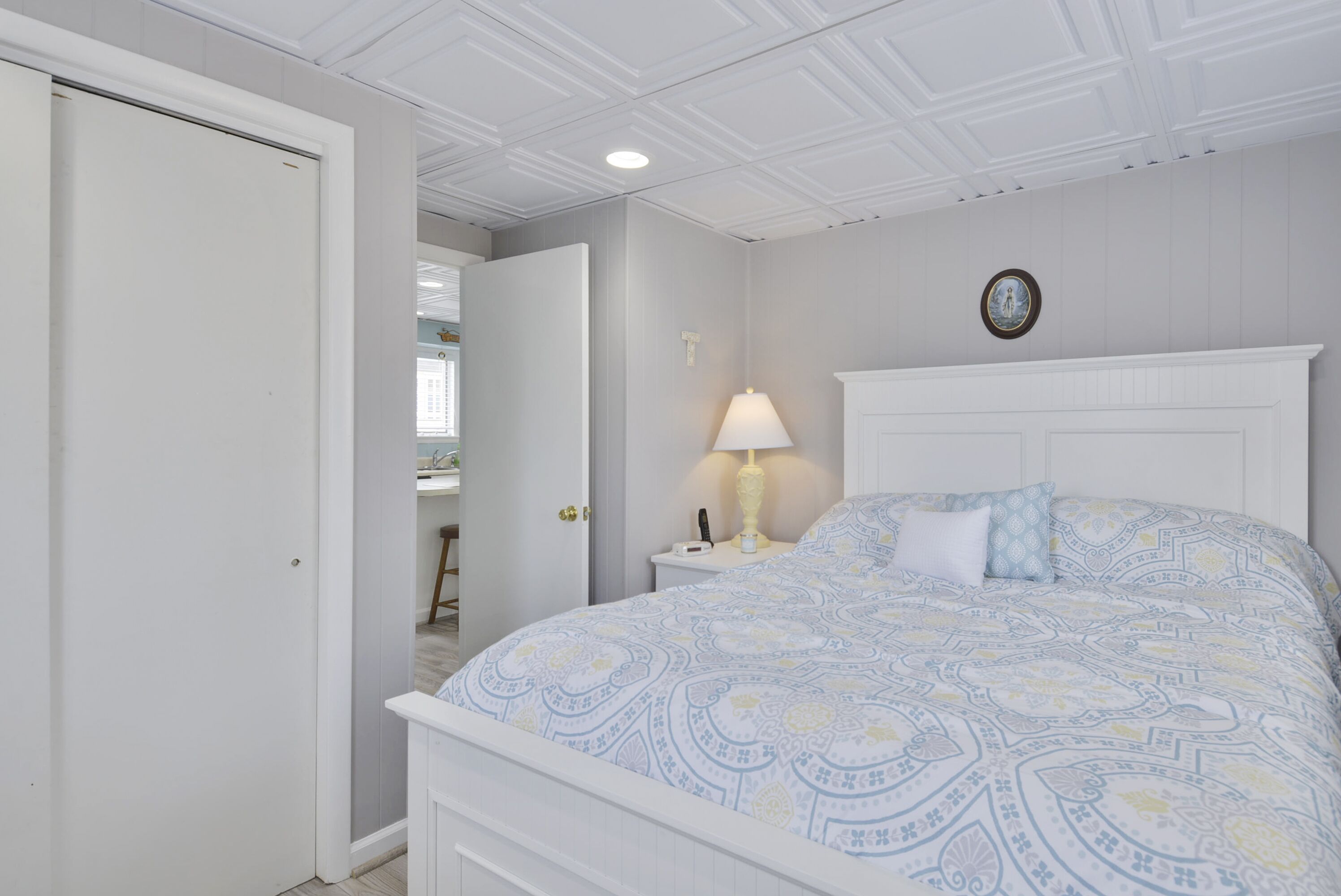 Een slaapkamer met een bed, nachtkastje met lamp, een wandklok en een deur naar een andere kamer.