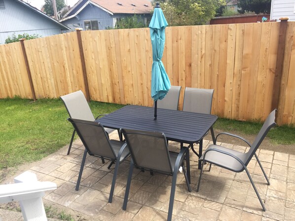 A patio with a metal table and four chairs, a blue umbrella, and a wooden fence in the background.