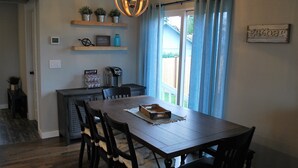 Un comedor con mesa de madera, sillas, una lámpara de araña y vistas a través de una ventana con cortinas azules.