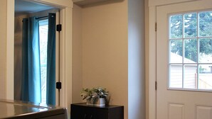 Un pasillo con una puerta, una ventana, un timbre, un felpudo y una pequeña mesa con una planta.