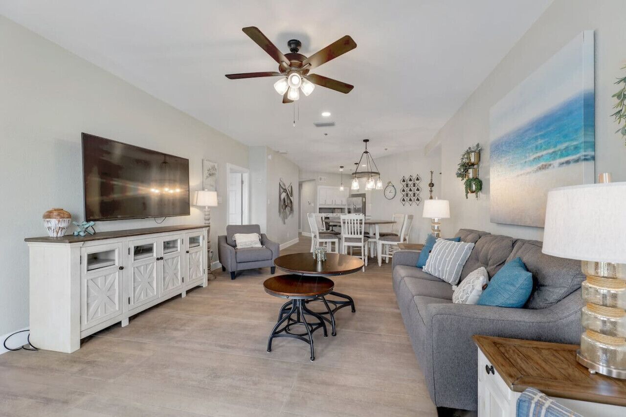 A spacious living room with a large flat-screen TV, a ceiling fan, a dining area with a table and chairs, and a gray sofa with blue cushions.