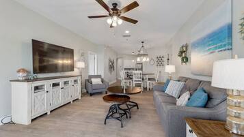 A spacious living room with a large flat-screen TV, a ceiling fan, a dining area with a table and chairs, and a gray sofa with blue cushions.