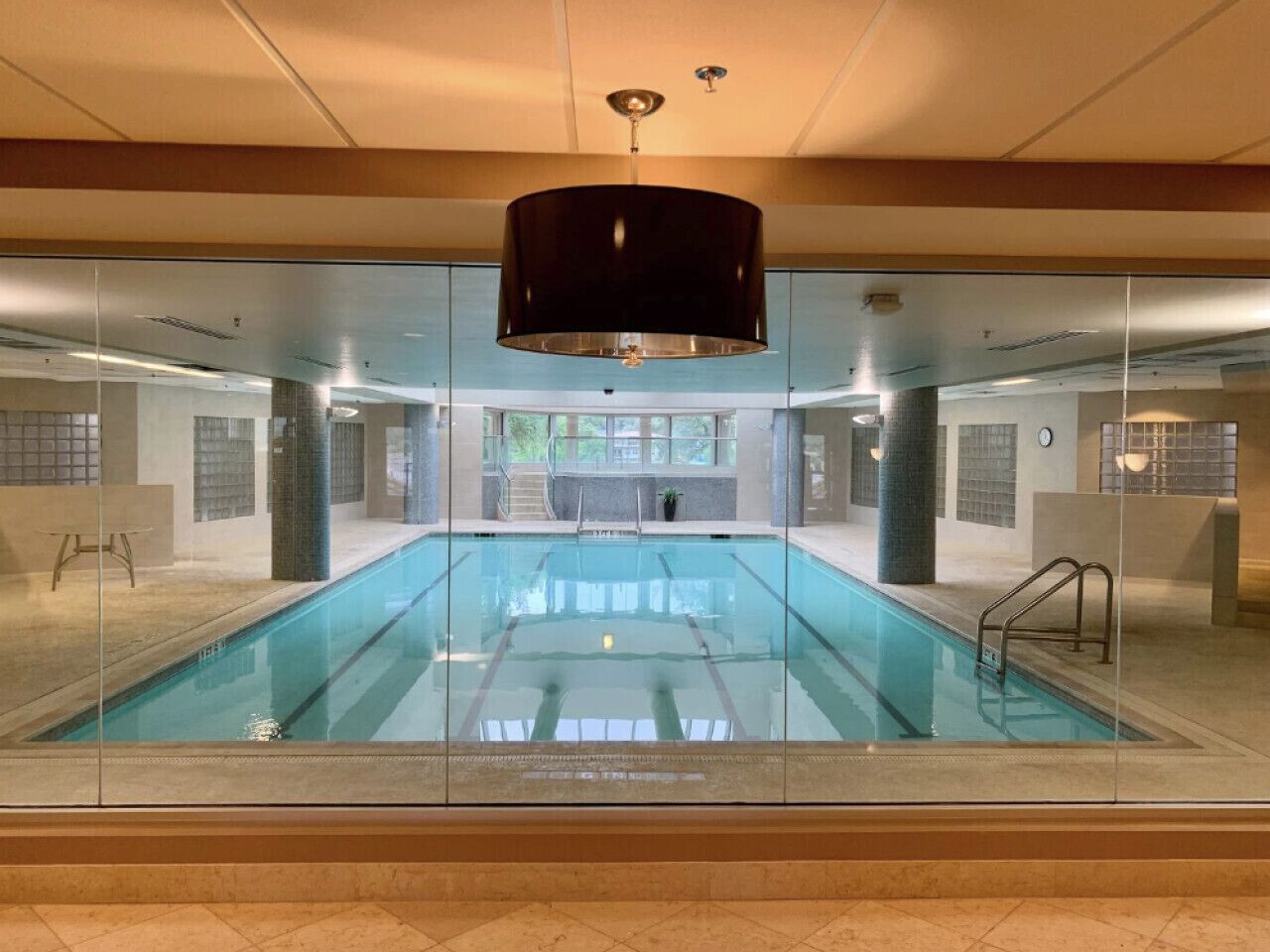 A large indoor swimming pool with clear glass walls, a black ceiling lamp, and a view of the outdoors through the glass.