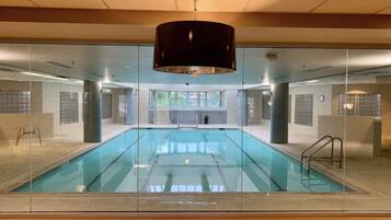 A large indoor swimming pool with clear glass walls, a black ceiling lamp, and a view of the outdoors through the glass.