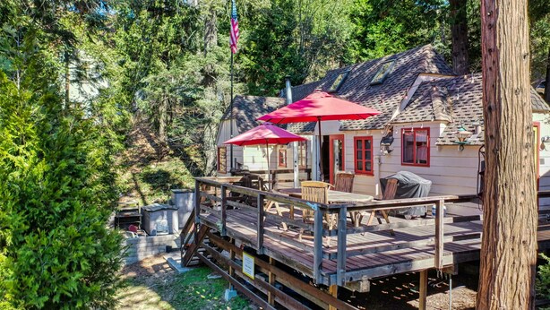 Eine Holzterrasse mit Sitzgelegenheiten im Freien, ein überdachter Bereich mit roten Sonnenschirmen und ein Haus mit Schornstein.