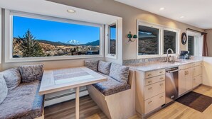 A modern kitchen with a dining area, large windows, and a view of a mountainous landscape.
