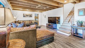 A spacious living room with a high ceiling, wooden beams, a sofa set, a flat-screen TV, and a staircase.