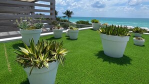 Un jardin sur le toit, avec du gazon synthétique et des plantes en pot, offrant une vue sur une plage et l’océan.