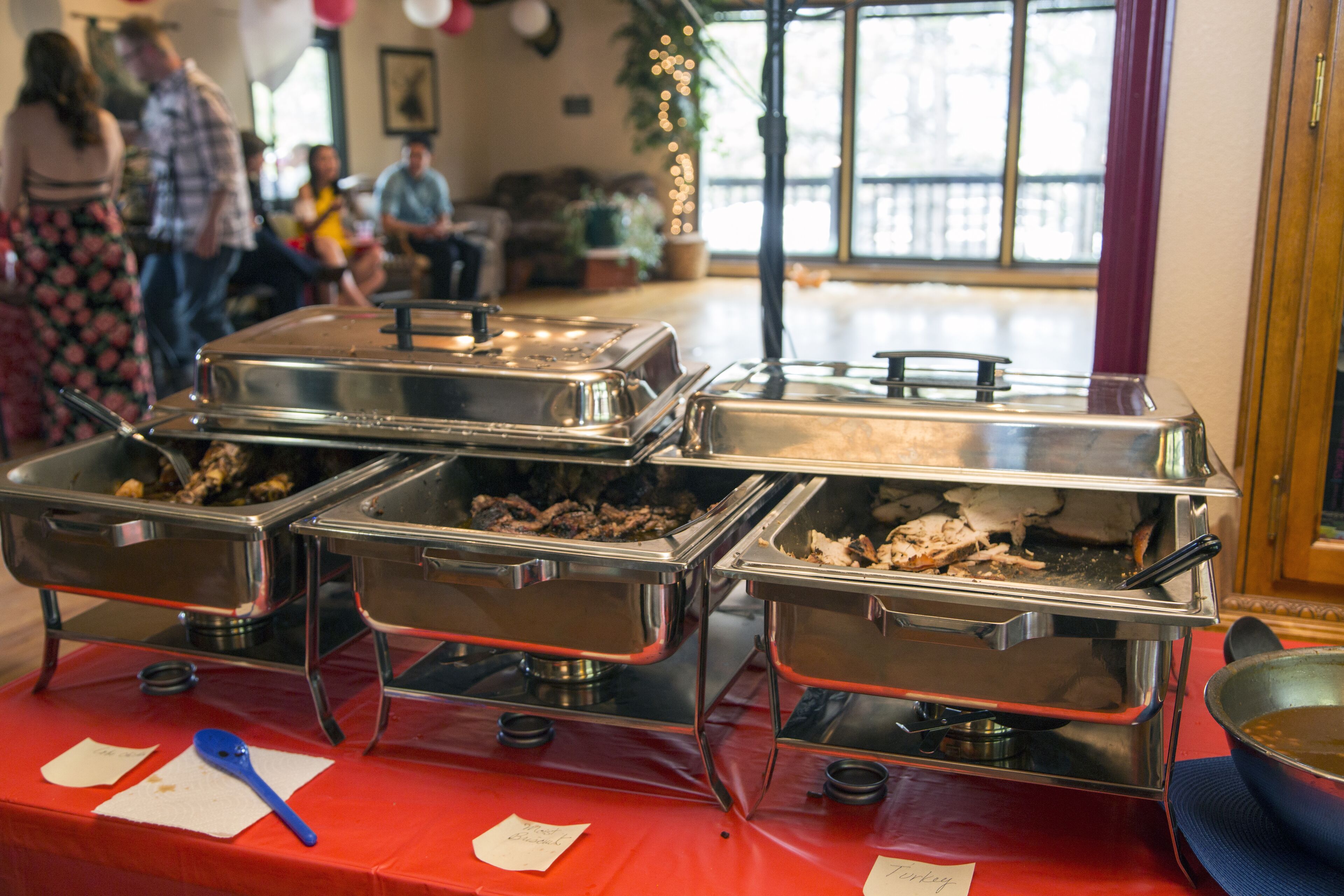 A buffet table with multiple stainless steel chafing dishes containing food, a red tablecloth, and a blue napkin.