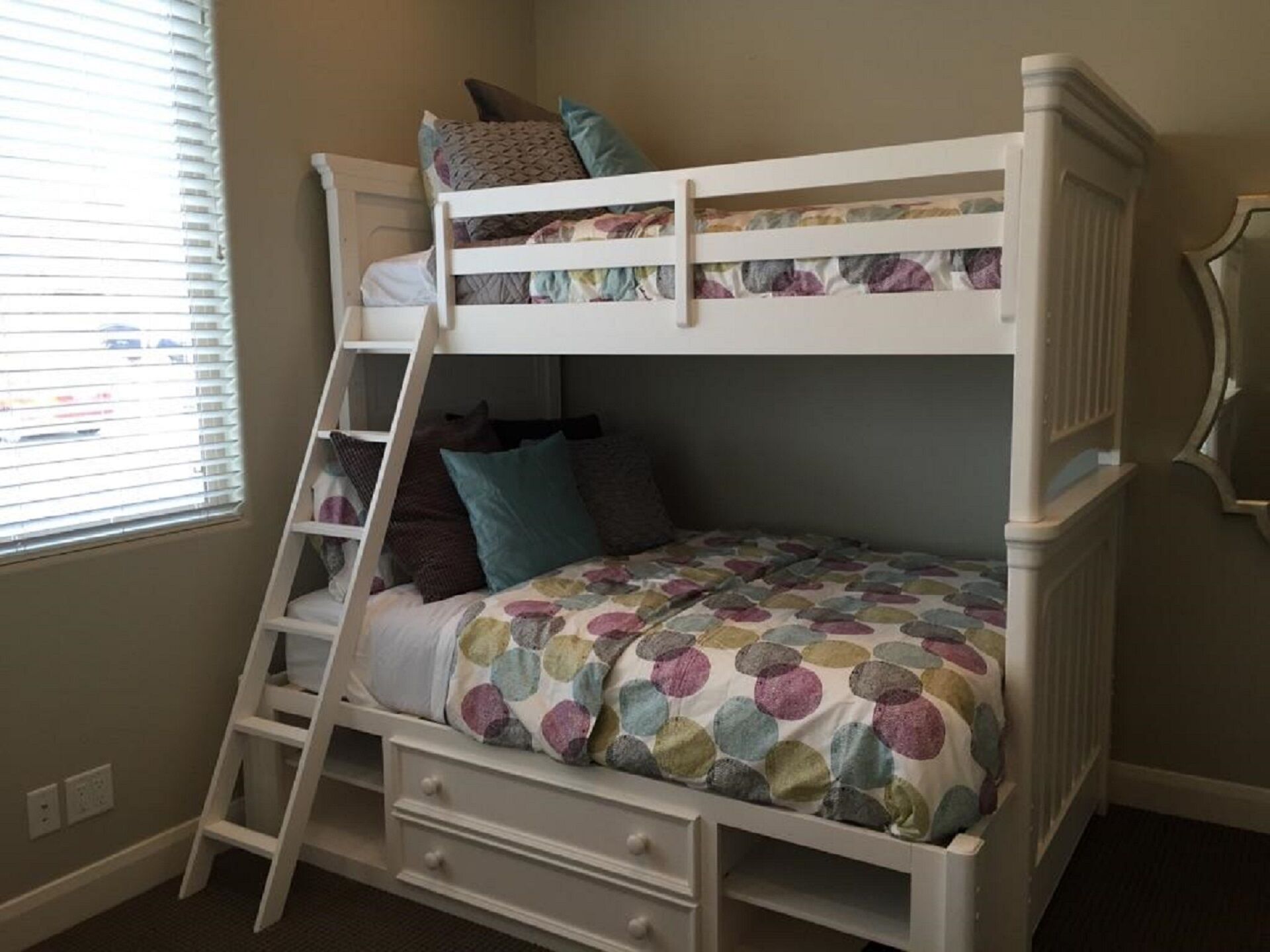 Een stapelbed met een ladder, een klein bureau en een raam met jaloezieën.