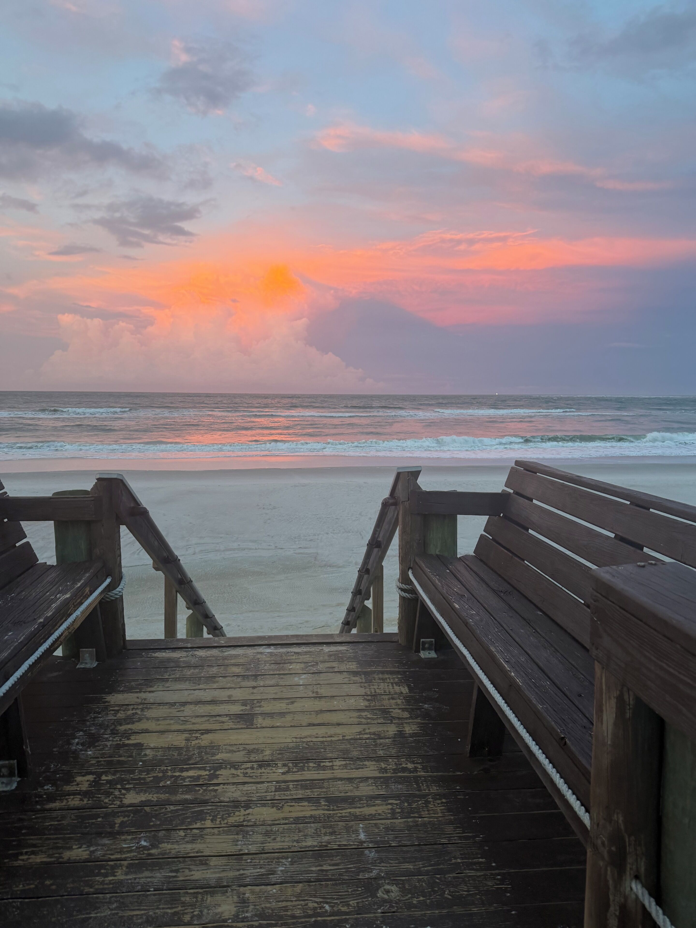 Ein Holzsteg führt zu einem Strand, im Hintergrund ein Sonnenuntergang.