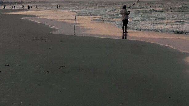 Une scène de plage avec une personne qui pêche, une mer calme et un ciel au coucher du soleil.