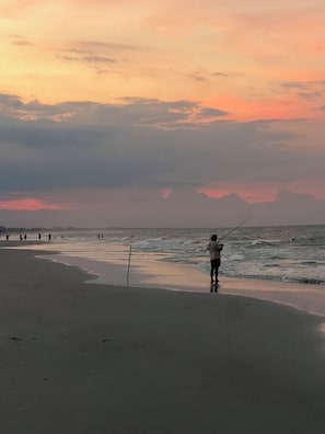 Une scène de plage avec une personne qui pêche, une mer calme et un ciel au coucher du soleil.