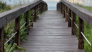 Un paseo marítimo de madera que conduce a la playa, bordeado por altas hierbas y barandillas de madera.