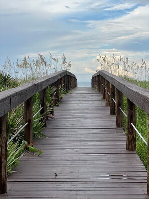 Une promenade en bois menant à la plage, bordée d’herbes hautes et de rampes de protection en bois.