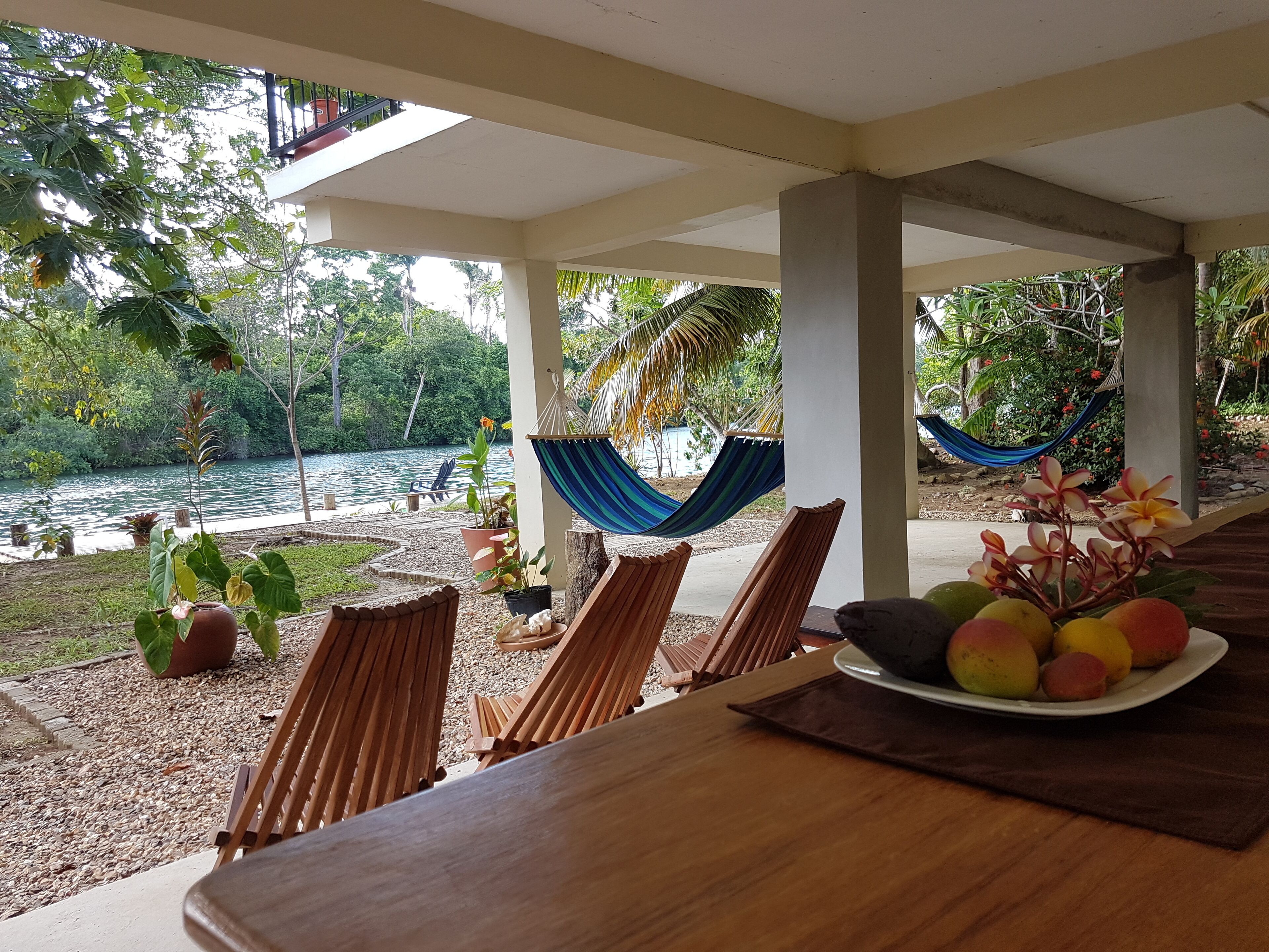 A wooden table with a plate of fruit, a hammock, and chairs under a covered area with a view of a lake and greenery.