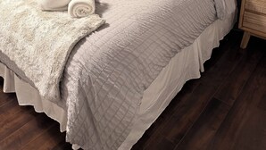 Un lit bien fait, avec une tête de lit grise, un plaid blanc moelleux et une serviette blanche roulée.