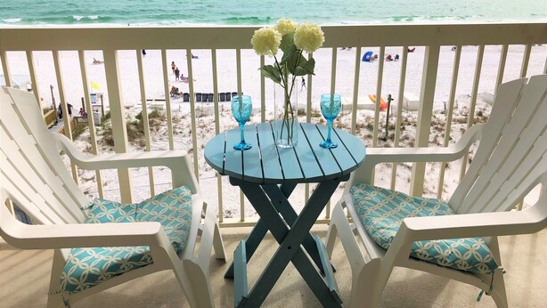 A balcony with two white chairs and a small table, overlooking a beach with people and beach equipment.