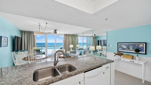 A modern kitchen with a granite countertop, stainless steel sink, and white cabinetry. There is a dining area with a table and chairs, and a large window offering a view of the ocean.
