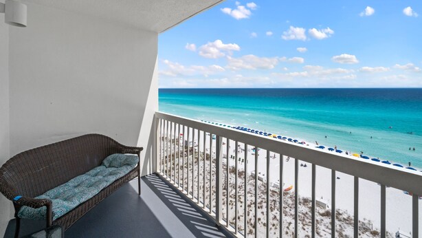 A balcony with a wicker sofa and a view of a beach with clear turquoise water and white sand.